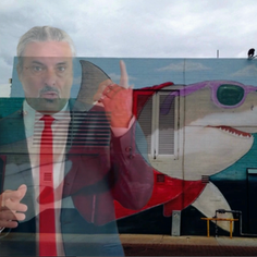 The King Teen, pointing upward with his left hand, superimposed on a photo of a building with a shark mural on it. 