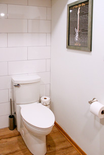 A new toilet rests on the finished wood floors with large format white stacked wall of tiles