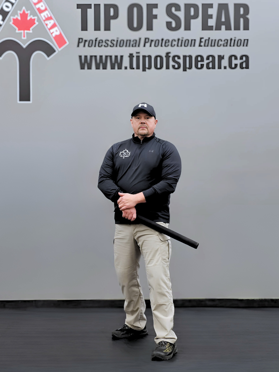 Man in black shirt and beige pants holds a baton. Background shows "TIP OF SPEAR" and website text. Serious expression, gray backdrop.