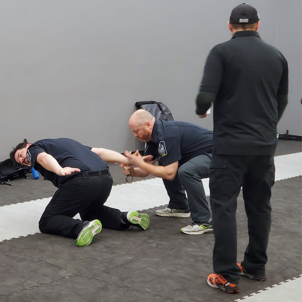 Three men in a training room; one on the floor, one kneeling using handcuffs, the third standing. Neutral colors dominate the scene.