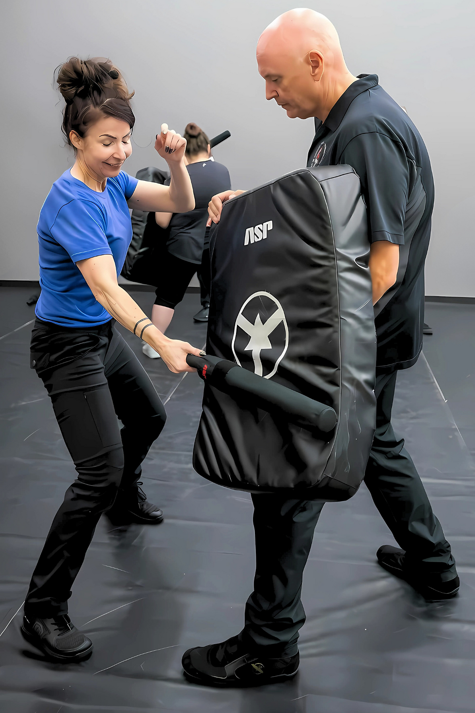 Woman in blue shirt practices self-defense with padded stick against a man holding a black shield. Indoor setting, focused expressions.