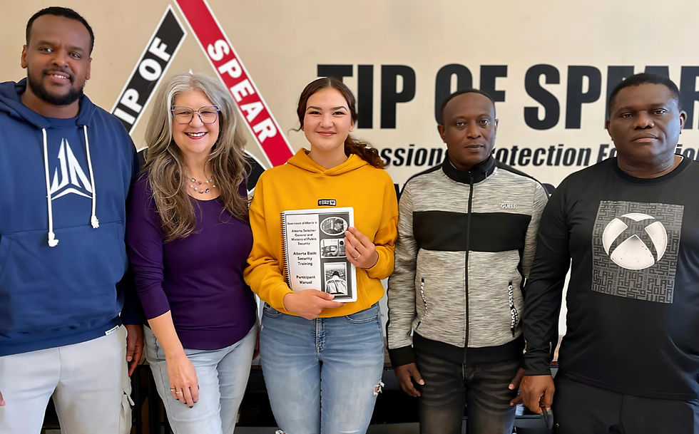 Five people stand smiling in front of a "Tip of Spear" sign. One holds a security training manual. Casual attire, cheerful mood.