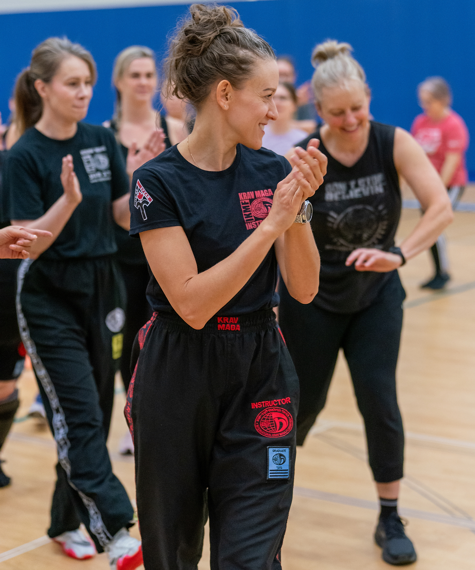 Women in a gym clapping during a class. They're wearing black athletic gear with red "Krav Maga" text. Blue wall in the background.