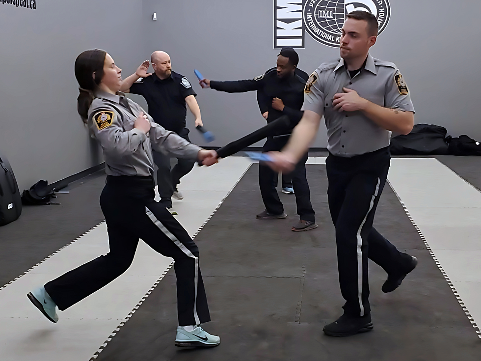 People in uniform practice self-defense moves in a gray room with IKMF logo. Two hold blue pads. Dynamic stance, focused mood.