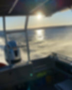 Boat speeding on lake as the sun is starting to set, sun glistening on water. Visible boat motor, splashing waves, and clear blue sky in background.