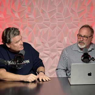 Rick Williams and Lonnie Spivak engaged in a podcast discussion, surrounded by a modern geometric backdrop illuminated with red lighting, with microphones and a laptop on the table.