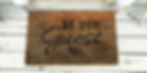 Weathered door mat on a wooden porch that reads Be Our Guest