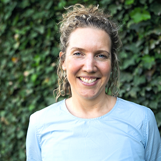 Ulla-Marie i ljusblå blus och uppsatt hår ståendes ute framför en vägg av gröna blad