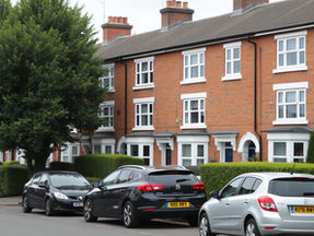 Cars parked on a residential street lined with red-brick houses and a large green tree. License plates visible, mood is calm and suburban.