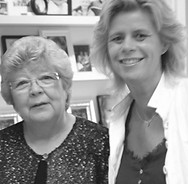 Oudere vrouw met bril en jongere vrouw tegen een muur met plankjes met foto's