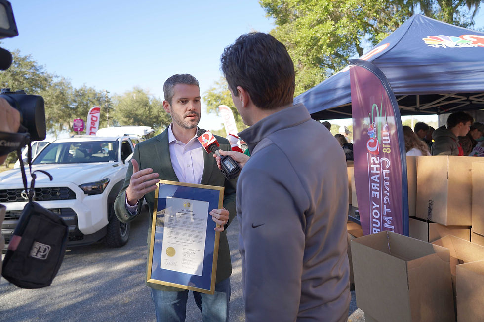 County commisioner framed certificate while being interviewed outdoors. A reporter with a microphone listens. Background features a car, tents, and banners.