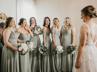 A bride in a white dress smiles at six bridesmaids in green dresses, each holding flowers, after a fun day of bridesmaid dress shopping