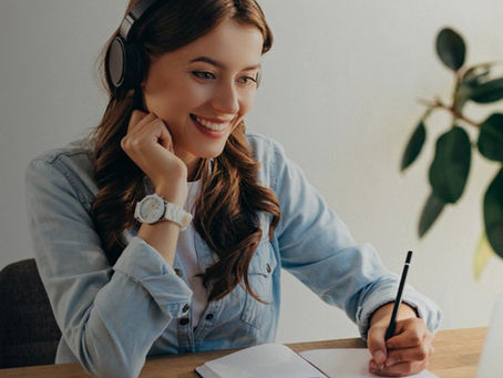 Lächelnde Frau mit Headset und Bleistift in der Hand sitzt vor einem Laptop und einem Notizbuch.