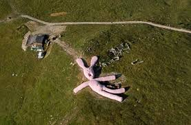 Aerial view of a large pink rabbit sculpture on a grassy hillside, near a path and small building, under a clear sky.