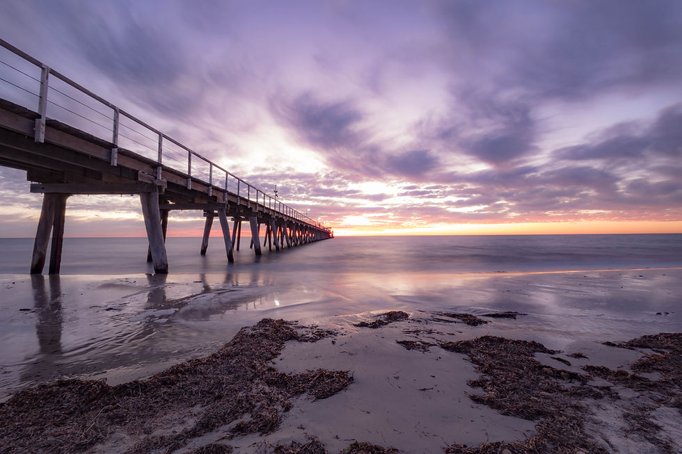 Sunset over a tranquil beach with a long pier stretching into the sea. Sky painted in purple and orange hues, creating a serene atmosphere.