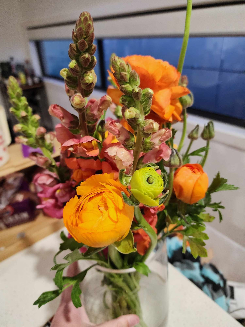 Bouquet of vibrant orange and pink flowers in a glass vase, held by a hand. Background shows a blurred indoor room and window.