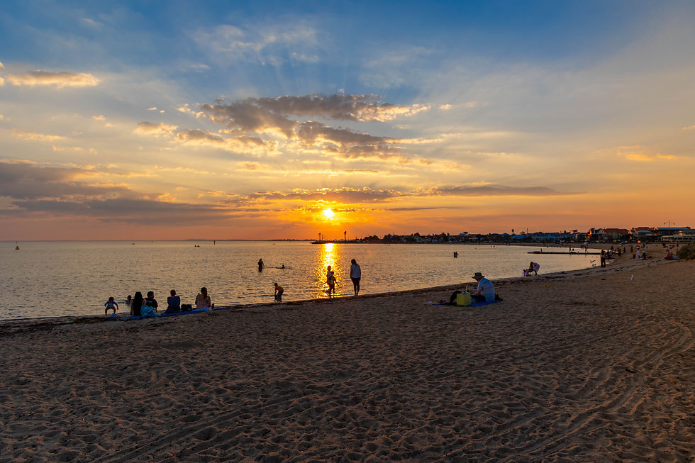 People relax on a sandy beach at sunset, with golden sky and calm water. Silhouettes of people standing in the water add to the serene mood.