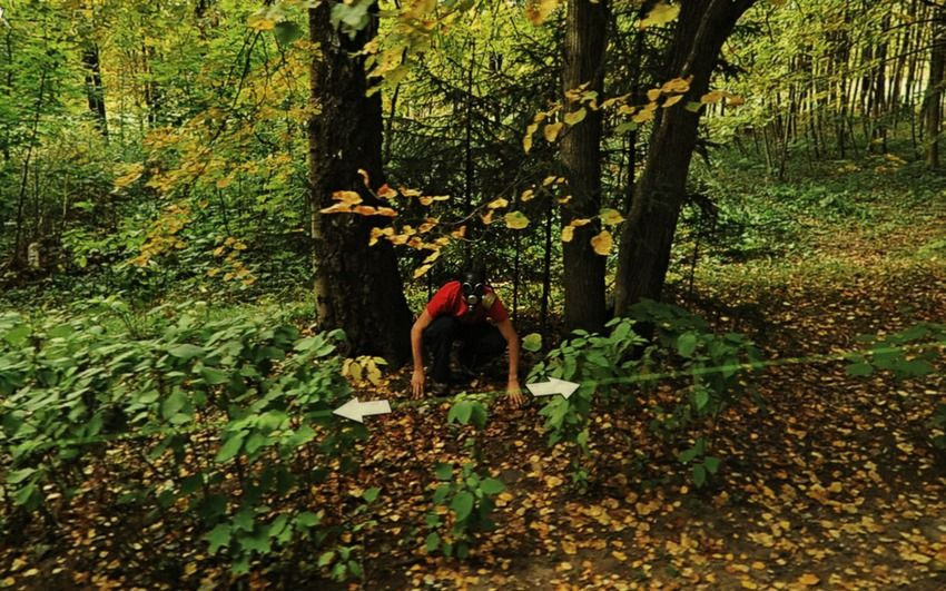 A person in a red shirt crouches under trees in a green forest. They are behind a white rope with arrows, surrounded by fallen leaves.