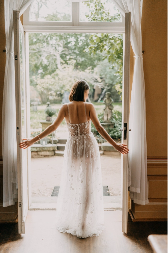 Moderne Braut im feinen Tüllkleid mit floraler Spitze öffnet französische Flügeltür – romantische Fine Art Hochzeitsinspiration mit mediterranem Flair
