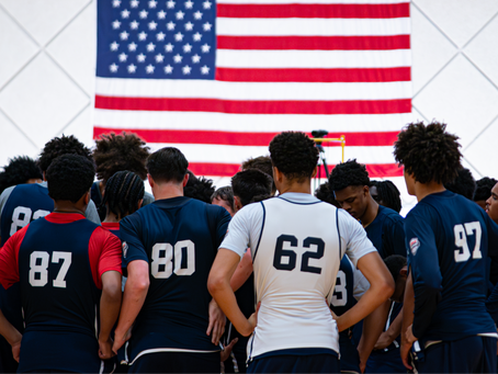 USA Basketball players in navy, red, and white practice jerseys huddle on the court beneath a large American flag hanging on the gym wall.
