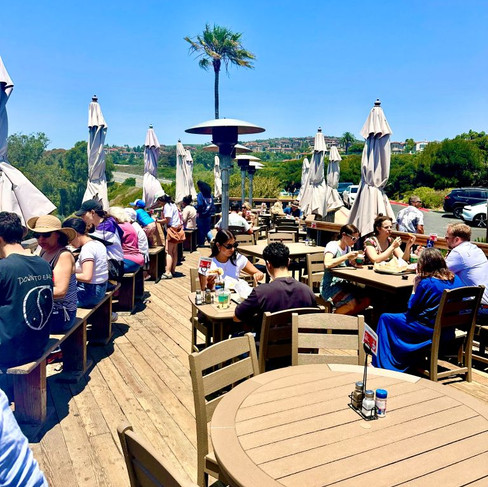 People sitting on at tables on the deck of an outdoor restaurant, eating and enjoying views on a sunny day.