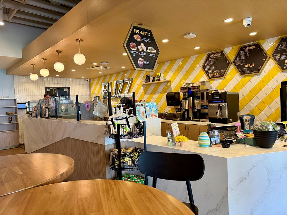 Cafe interior with yellow and white wall tiles, round light fixtures, and "BUZZ" text. Counter displays snacks and coffee machines. Cozy ambiance.