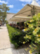 Outdoor cafe with people dining under white canopies. Green hedges, yellow flowers, and a sunny sky provide a lively, relaxed atmosphere.
