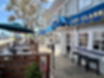 Outdoor café with blue awning and "EAT CLASSIC" sign. Empty tables and chairs under umbrellas. Tree, ocean, and road in background. Calm mood.