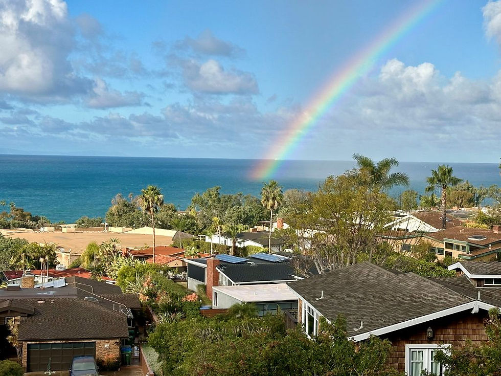 A rainbow stretches from a cloudy sky to the ocean, beyond the roofline of a neighborhood in Laguna Beach.