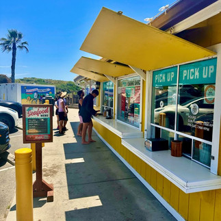 A line of people waiting to order at a yellow food shack with "pick up" windows next to a palm tree.