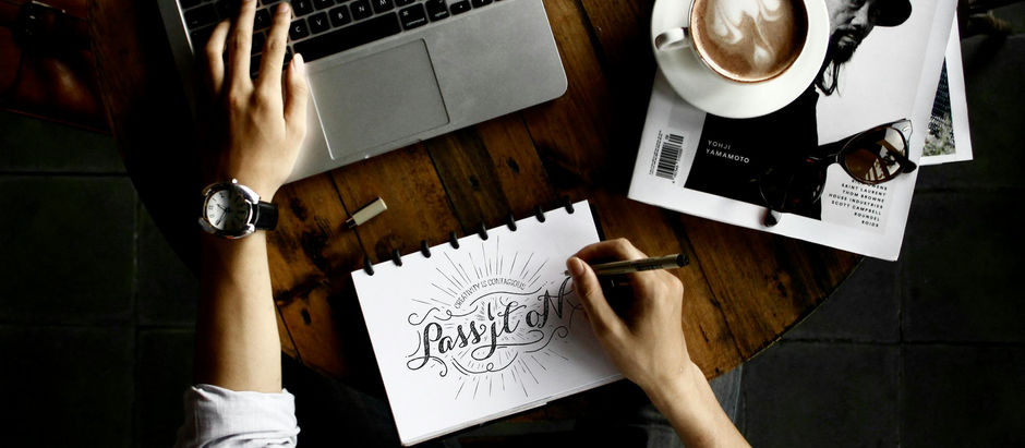 Person in denim shirt writing in a notebook on a wooden desk with a laptop, black book, and pen holder. Cozy, focused workspace.
