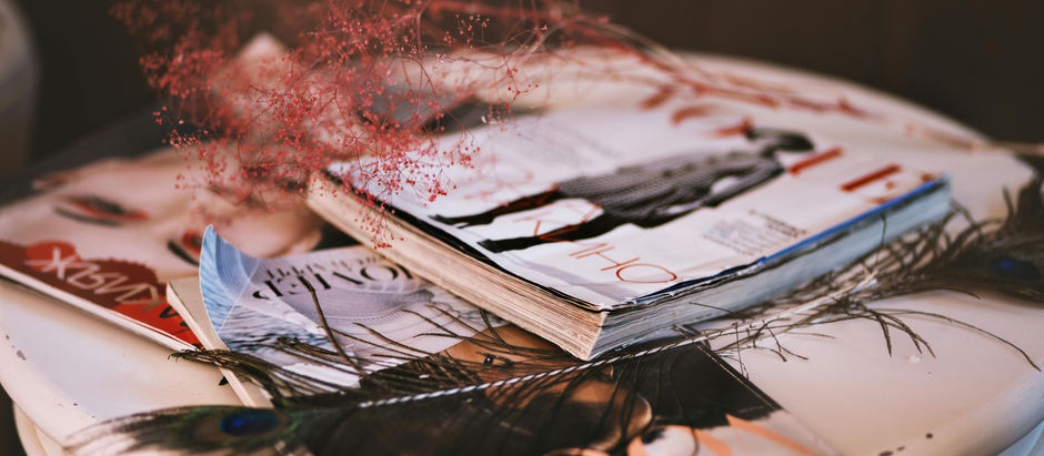 Magazines stacked on a white table with pink dried flowers and a peacock feather. Covers feature fashion photography and text. Cozy mood.
