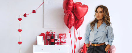 Michelle Herasme, Board-Certified Nurse Practitioner and founder of Herasme Beauty, standing in her Englewood, NJ studio decorated with red heart balloons and Valentine's Day gifts including wine, wrapped presents, and heart decorations
