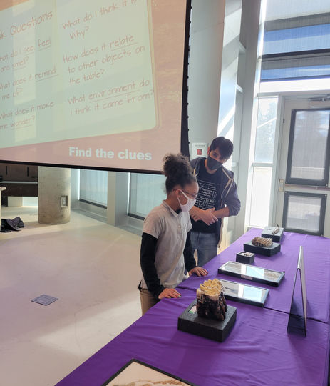 Students examining artifacts; "Find the clues" on screen