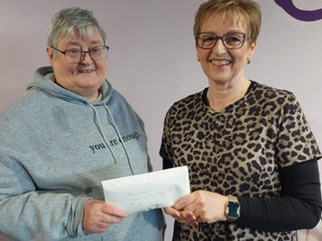 Two women stand side by side smiling at the camera, with the woman on the left wearing a grey hoodie that says "you are enough" and the woman on the right in a leopard print top, both holding a small white cheque between them.