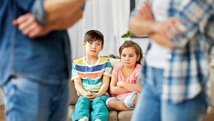 Confused and frustrated, two children sit on a couch as their parents debate parenting decisions.