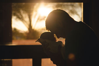 A man in silhouette, holding a baby in front of him, pressing his forehead to its forehead. Behind them the sun is setting through a window.