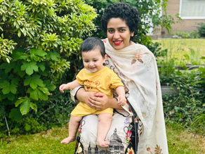 Tooba is a Pakistani woman wearing a flowing Pakistani dress and scarf. She is sitting in a garden with her young child on her lap. She is smiling and happy.