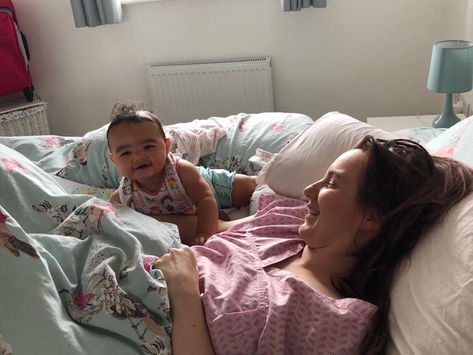 Kristie is lying in bed with her baby daughter crawling over the cover near her chest. Kristie is smiling but looks tired