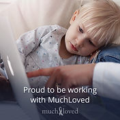 A small boy with blonde hair leans against a sofa cushion looking at a laptop screen. An adult's hand is pointing to the screen