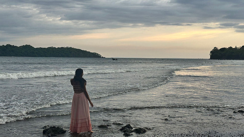 Woman in a pink skirt stands on a beach, gazing at the ocean under a cloudy sky. Islands and a small boat are visible on the horizon.