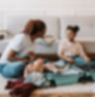Mother and daughter smile while packing clothes into a mint suitcase on the floor of a bright living room.