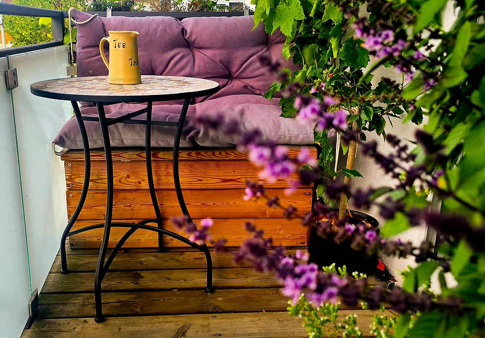 Bild von Stefanie Kießlings Balkon. Es ist eine Holztruhe mit einer gemütlichen, lila Polsterauflage zu sehen. Davor steht ein kleiner Bistrotisch mit Moasiksteinen und Metallgestell. Auf dem Tisch steht eine Kann mit der Aufschrift Tee. Es sieht so aus, als würde man zwischen Blüten vom Strauchbasilikum einen kleinen Blick erhaschen. Der Balkon hat einen Holzboden und eine Umrandung aus weißem Milchglas.