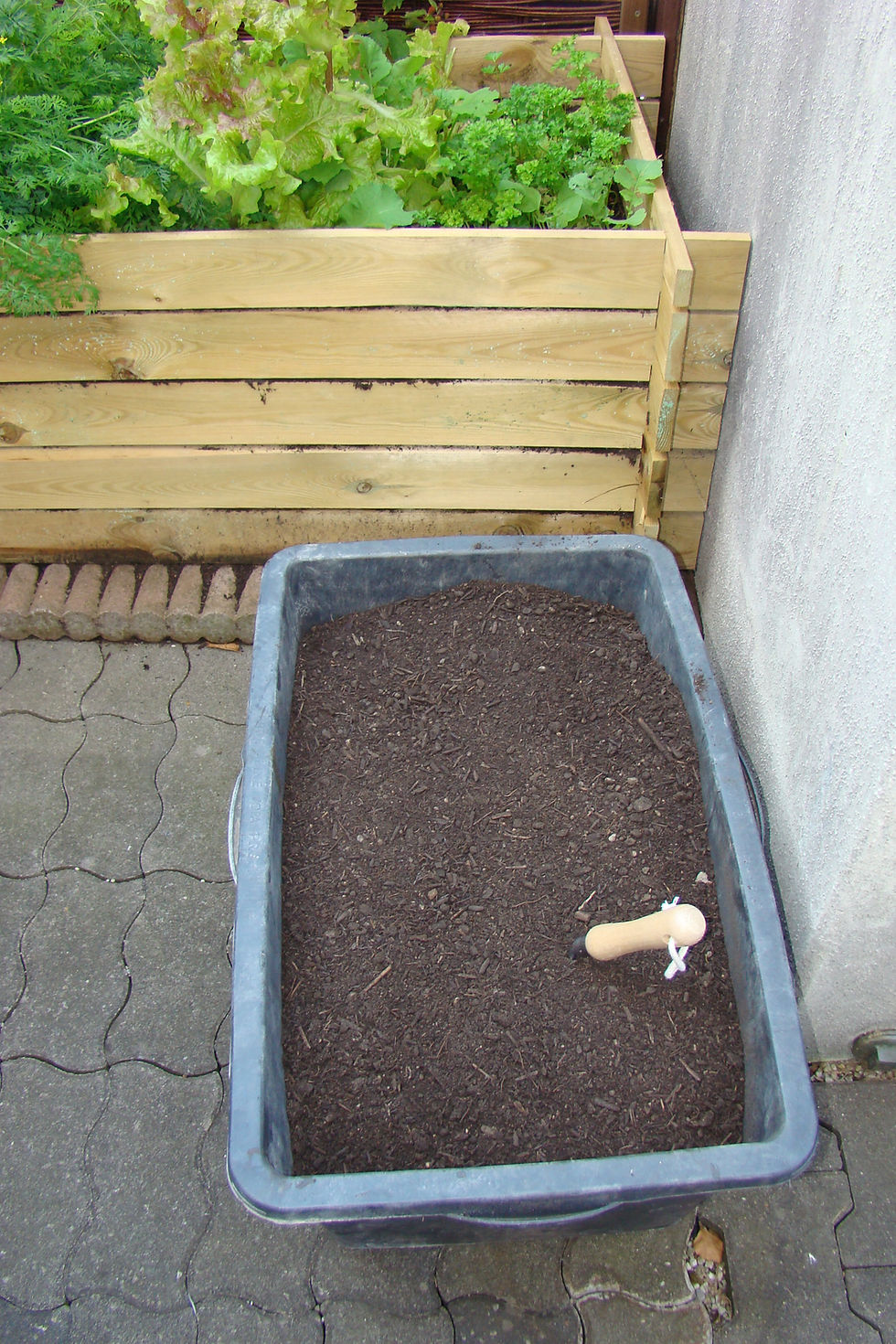 schwarze Bauwanne mit Erde gefüllt. Darin steckt eine kleine Schaufel mit einem Holzgriff. Im Hintergrund ist ein Hochbeet aus Holz mit Salaten zu sehen