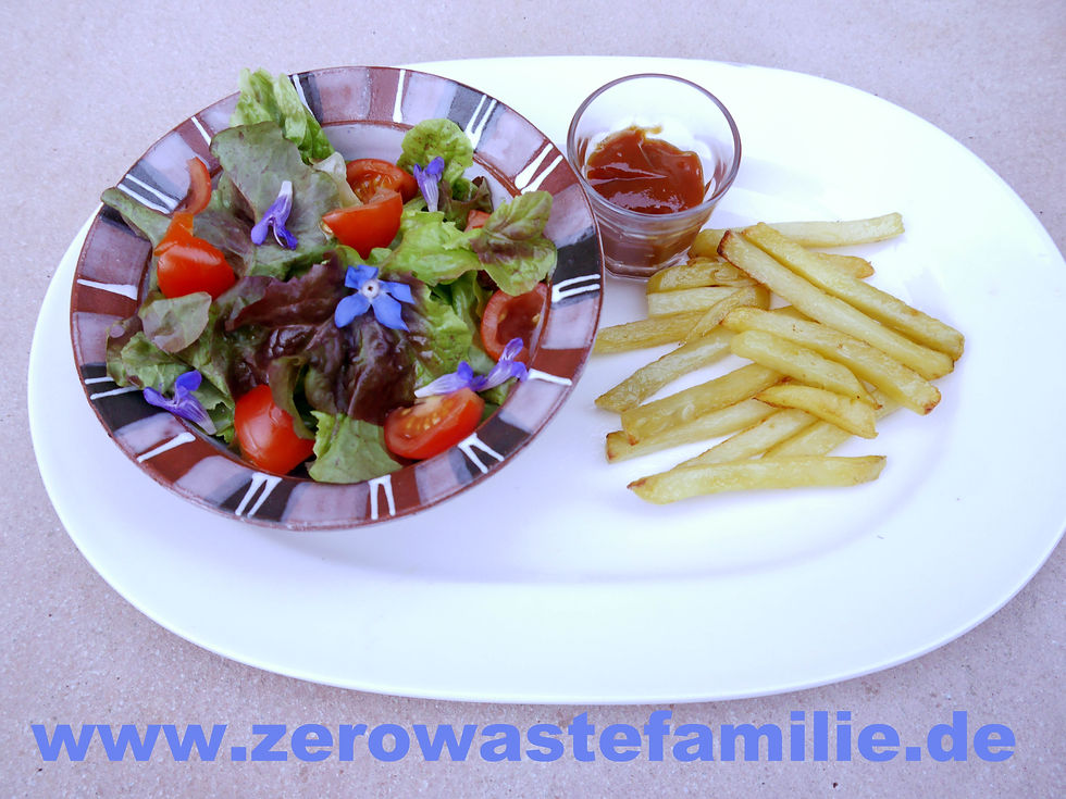 Pommes auf einem großen weißen Platte aus Porzellan. Daneben eine kleine Glasschüssel mit Ketchup und eine Keramik Schüssel mit bunten Salat und blauen Blüten
