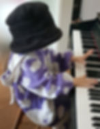 kleines Kind sitzt am Klavier in einem lila Milka-Kuh-Kostüm und einem riesigem schwarzen Hut, der komplett den Kopf verdeckt