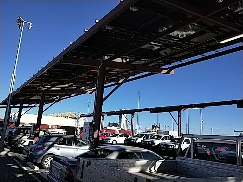 Cars parked under solar panel canopies in a lot. Clear blue sky and utility poles in the background. Bright, sunny day.