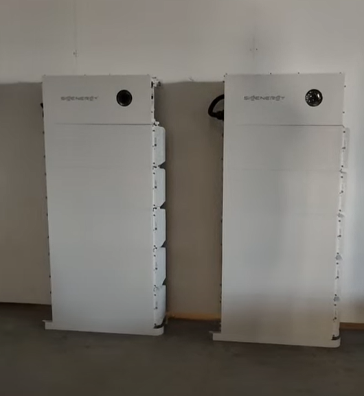 Two large white battery units stand against a plain wall. The word "SIGENERGY" is visible on both. The setting is minimal and industrial.