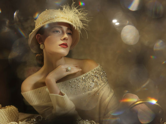 Model in cream cashmere sweater with crystal beading and ornate feathered headpiece, photographed with dramatic golden lighting