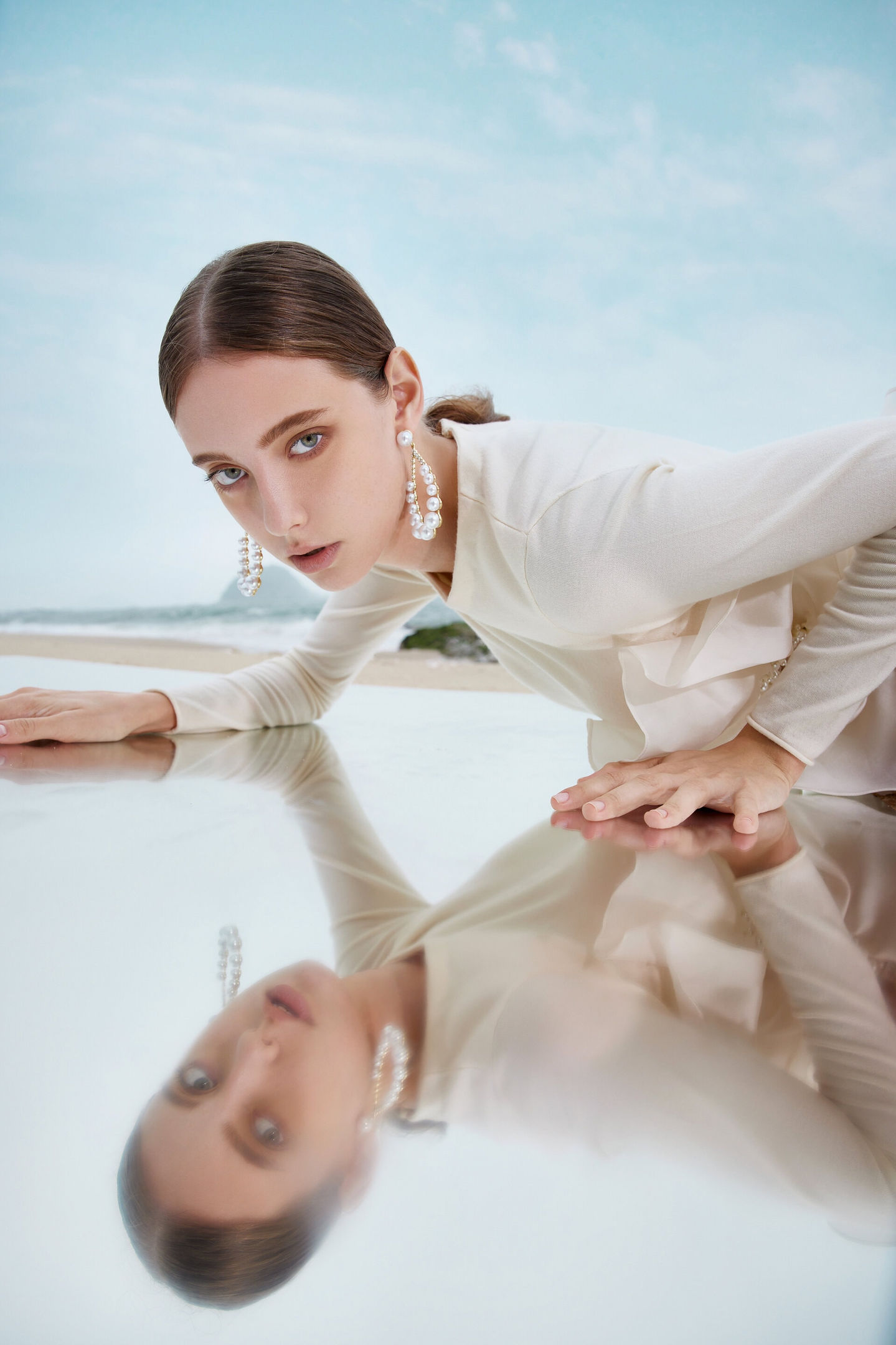 Model wearing a refined Cashmere Song gown with pearl embellishments, captured in a reflective pose on the beach